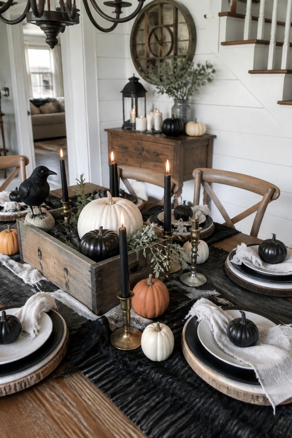 Dining table with a wooden box centerpiece holding black and white pumpkins, black candles, and a black crow figure, surrounded by layered black and white plates and small pumpkins on a dark runner.