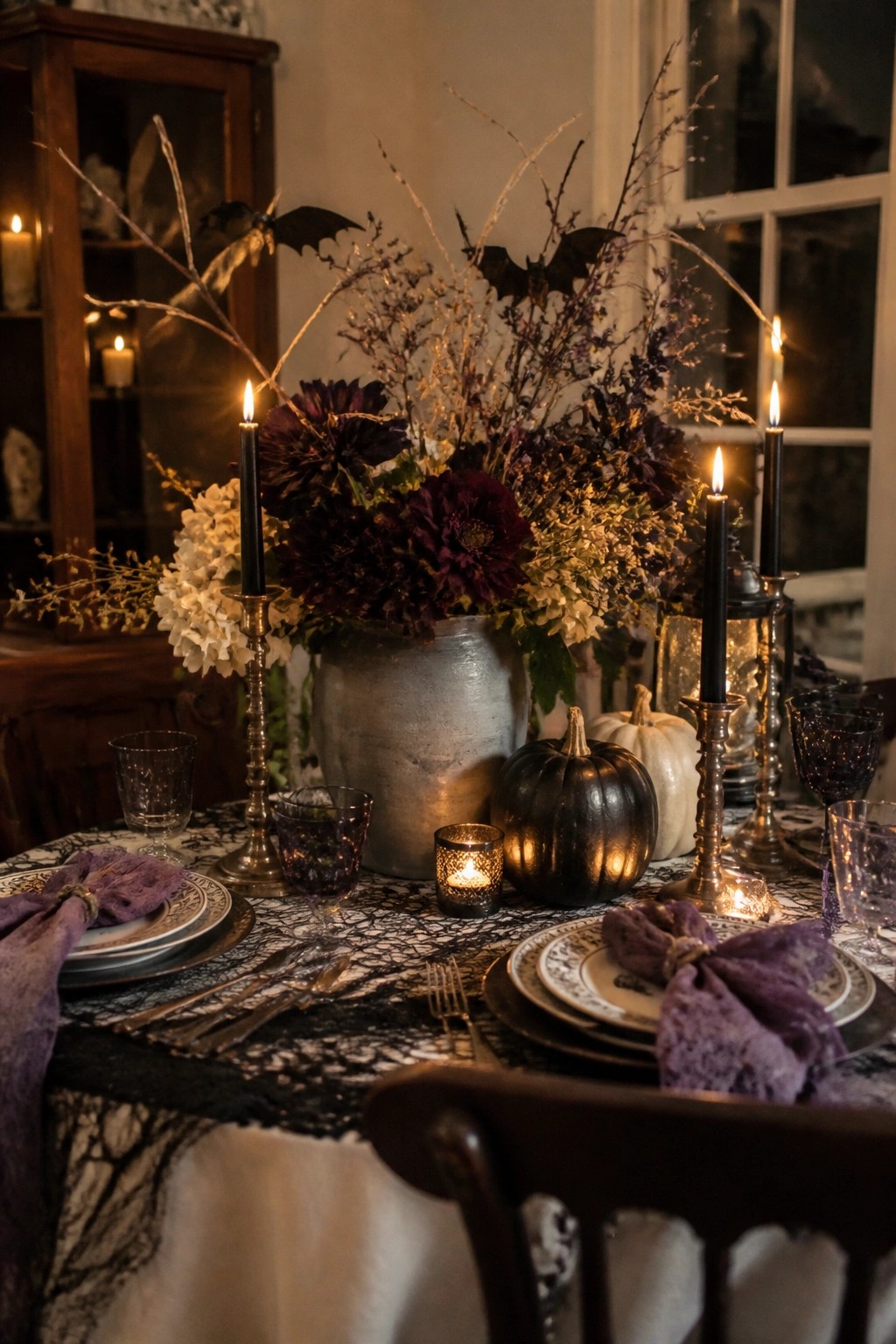 Dining table set for Halloween with a large floral centerpiece featuring dark purple flowers, dried branches, and black bat figures, surrounded by black candles, pumpkins, and purple napkins on a spiderweb lace runner.