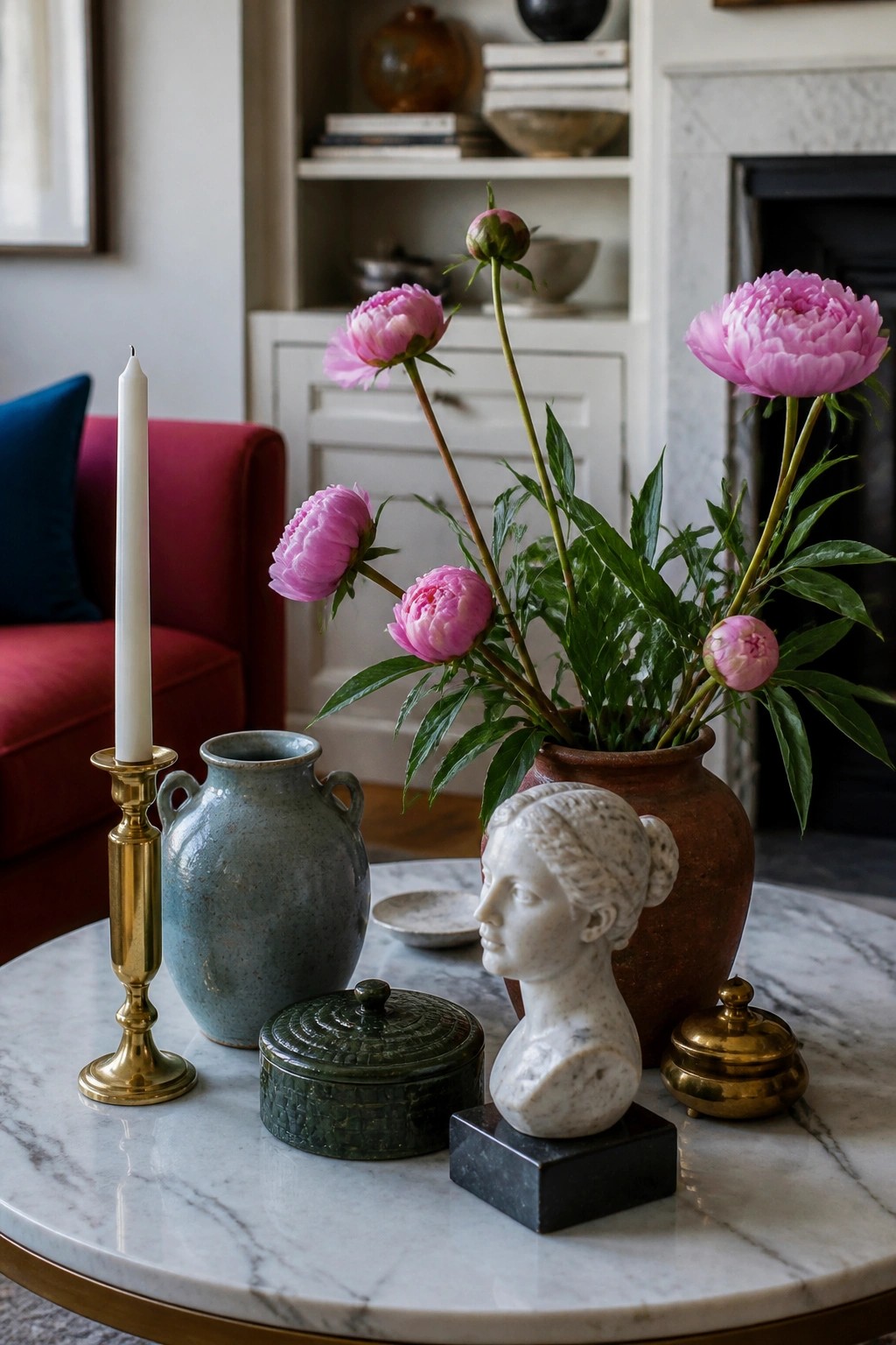Marble coffee table with a white stone bust, brass candle holder, green lidded box, and a terracotta vase filled with pink peonies.