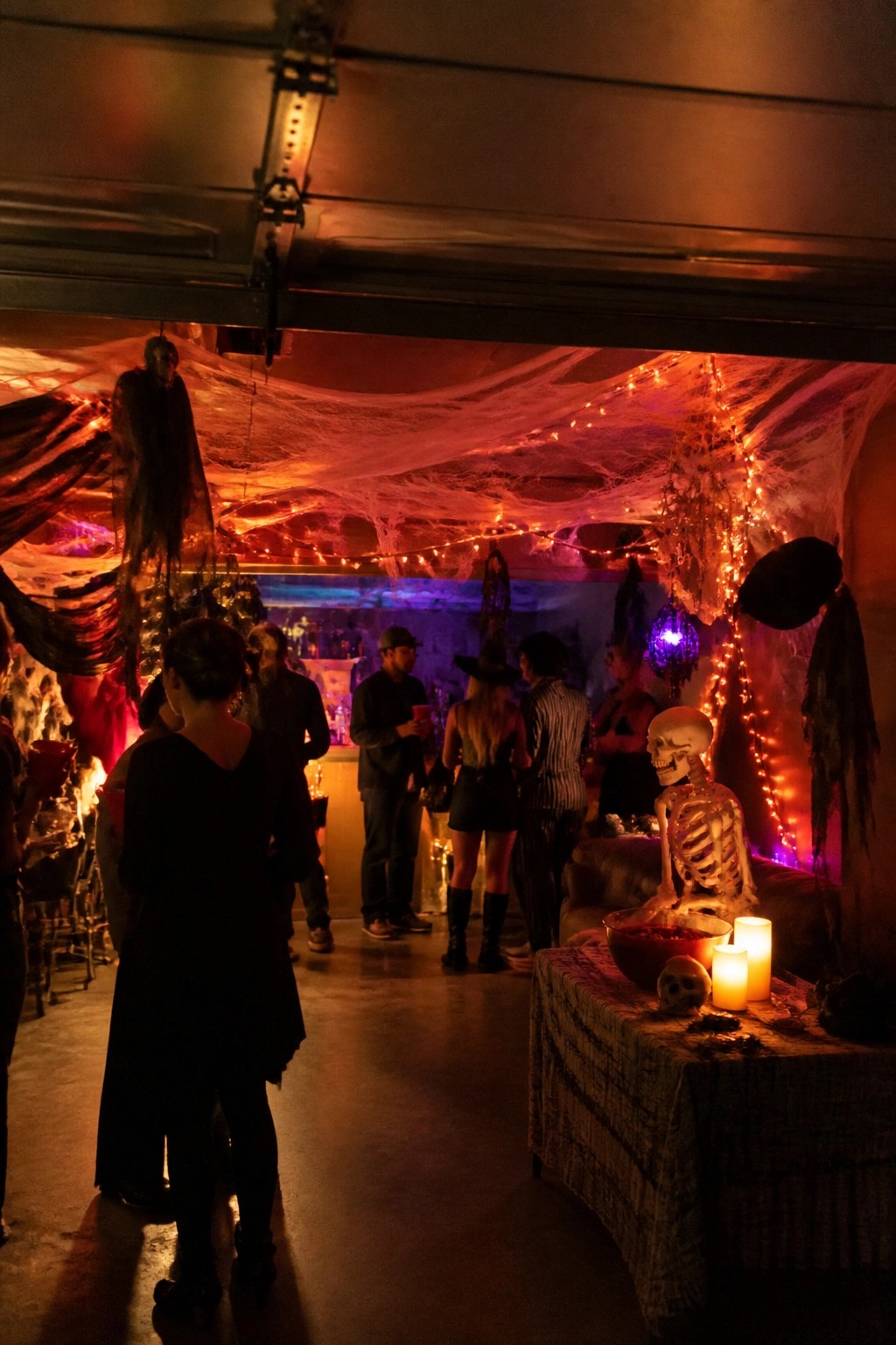 Garage party space with fake spiderwebs covering the ceiling, string lights woven through them, and a skeleton seated on a couch.