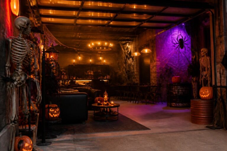 Industrial garage space with multiple hanging ghost figures suspended from the ceiling, skeletons on the walls, black leather seating, and glowing jack o lanterns arranged for a Halloween party.