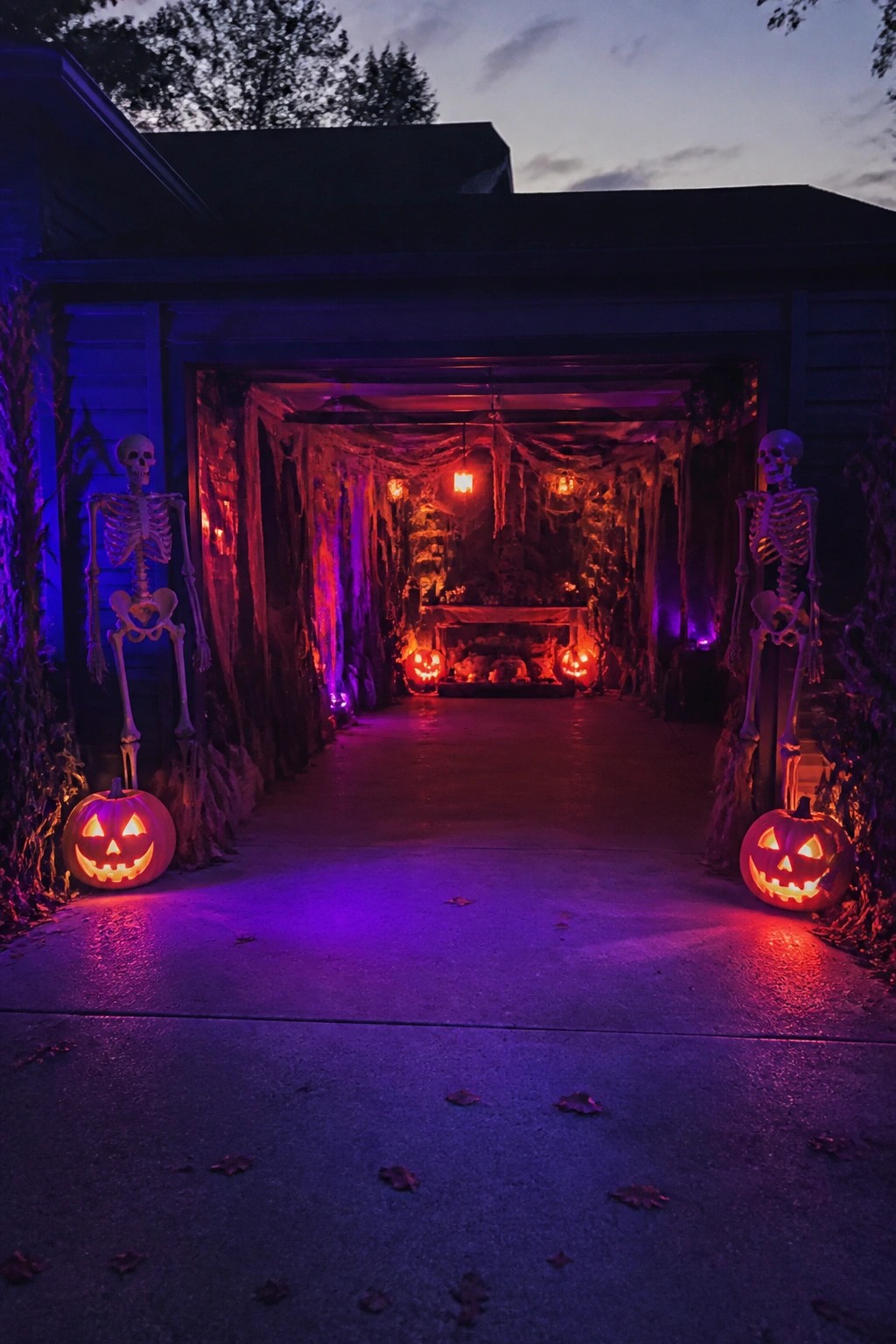 Garage entrance lined with two standing skeletons, jack-o'-lanterns at the base, and heavy cobwebs and fabric draped throughout the interior.
