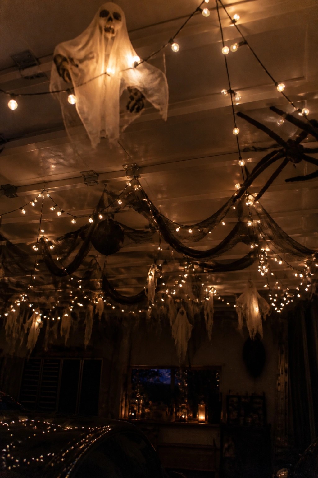 Garage interior with multiple white fabric ghosts hanging from the ceiling among string lights, black webbing, and large spider props.
