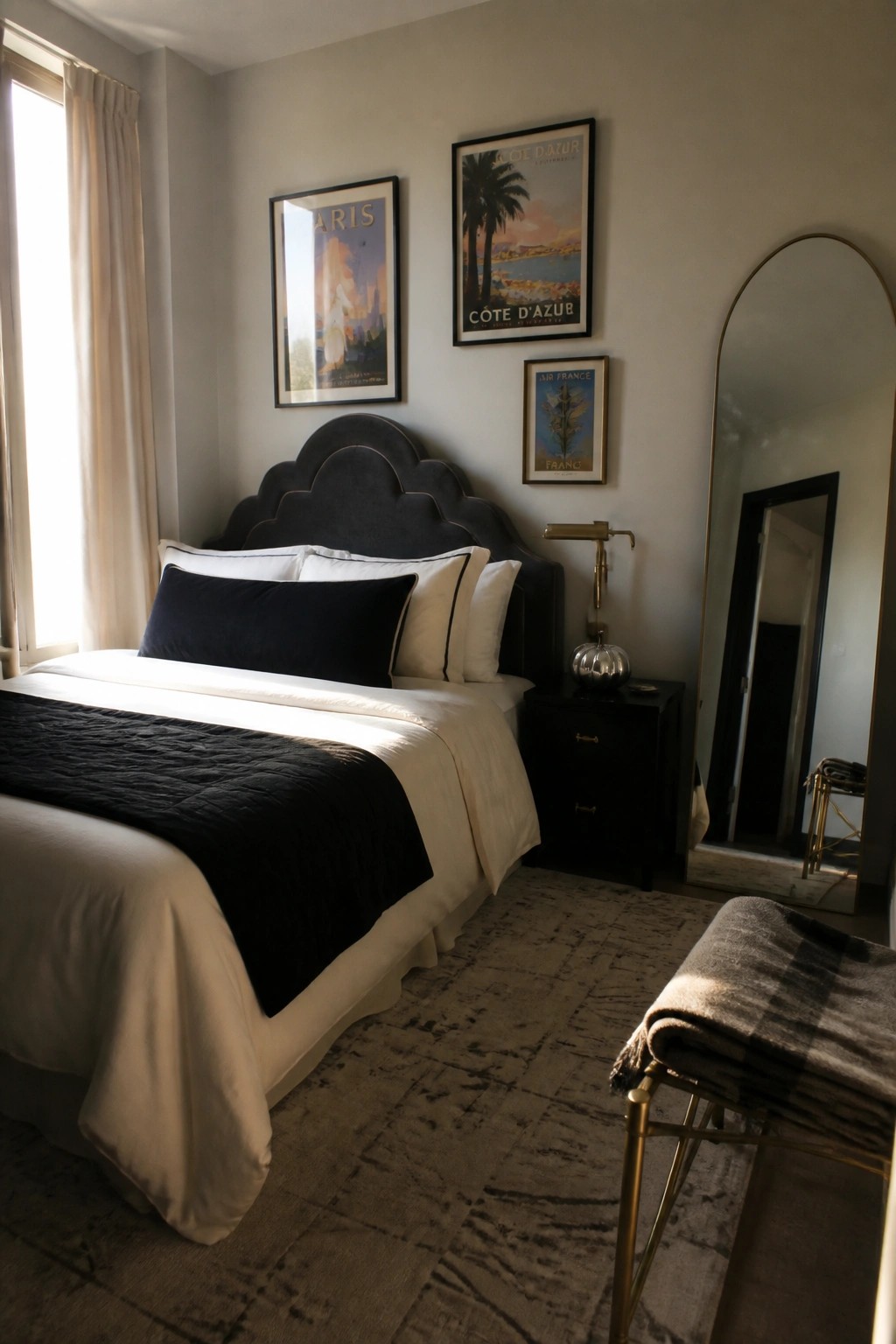 A bedroom with a dark scalloped headboard, black and white bedding, a black nightstand, and three framed travel posters on a light wall.