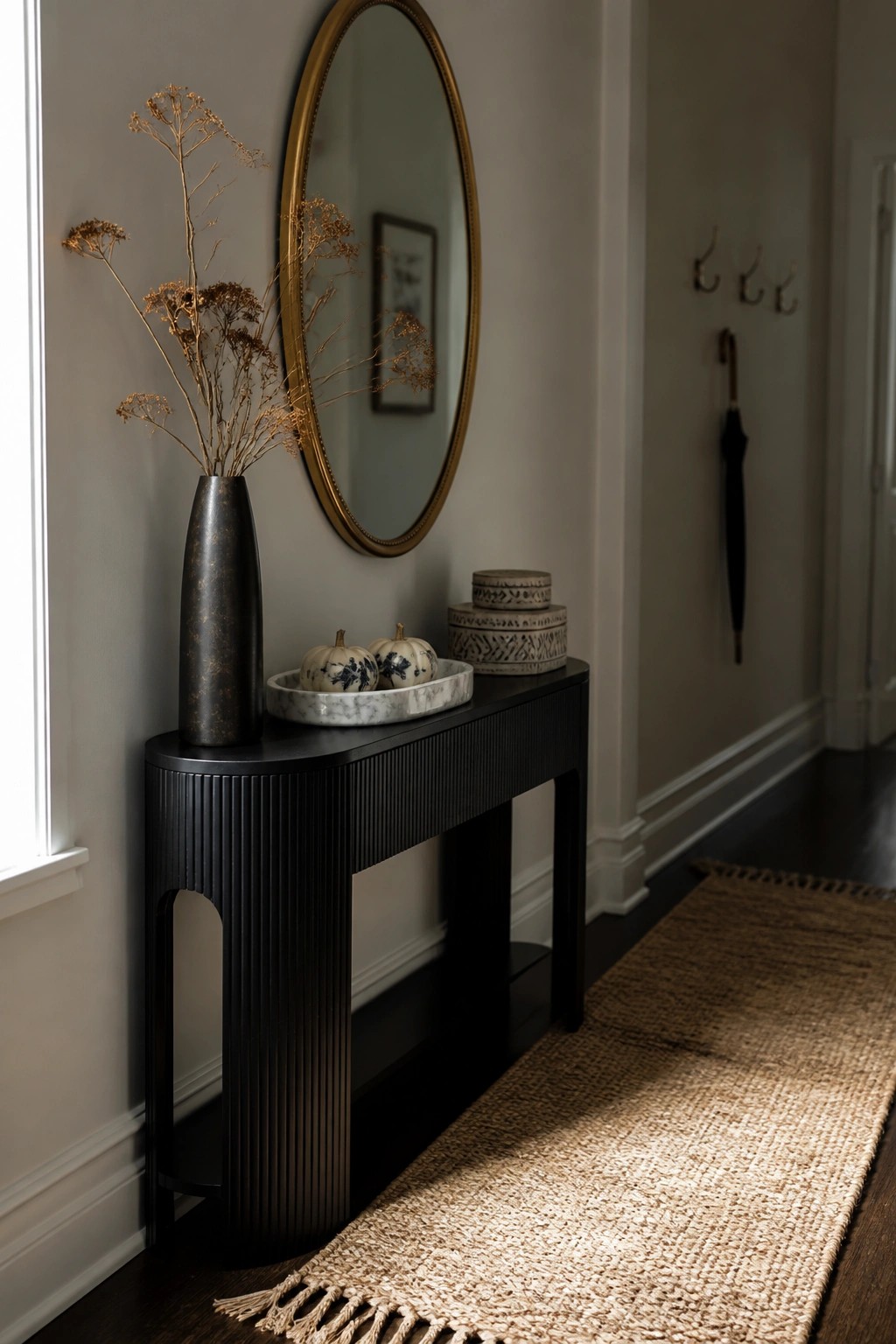 A black ribbed console table against a light wall holds a tall dark vase with dried flowers and a marble tray containing two small white pumpkins, with a round gold mirror above and a woven jute runner on the dark wood floor.