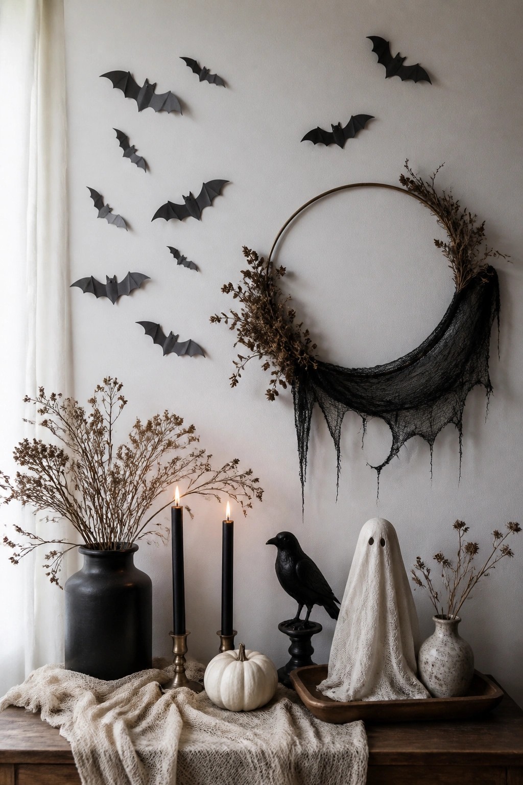 White wall decorated with multiple black paper bats in flight above a wooden table holding candles, a small white pumpkin, a black crow figurine, and a ghost figure.