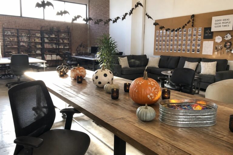 An office conference table with black and white painted pumpkins, a bowl of candy, and small candles, plus black bat garlands hung along the brick wall behind.
