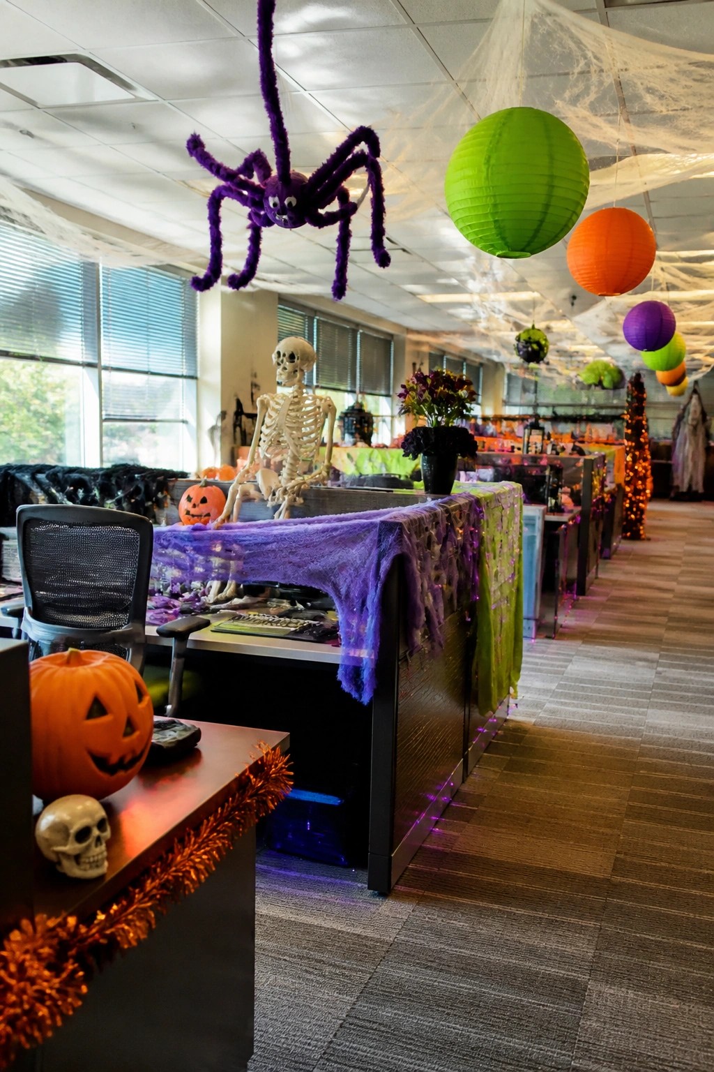 Office space with cubicles decorated for Halloween, featuring a large purple spider hanging from the ceiling with fake webs, a skeleton seated at a desk, pumpkins, and colorful paper lanterns.