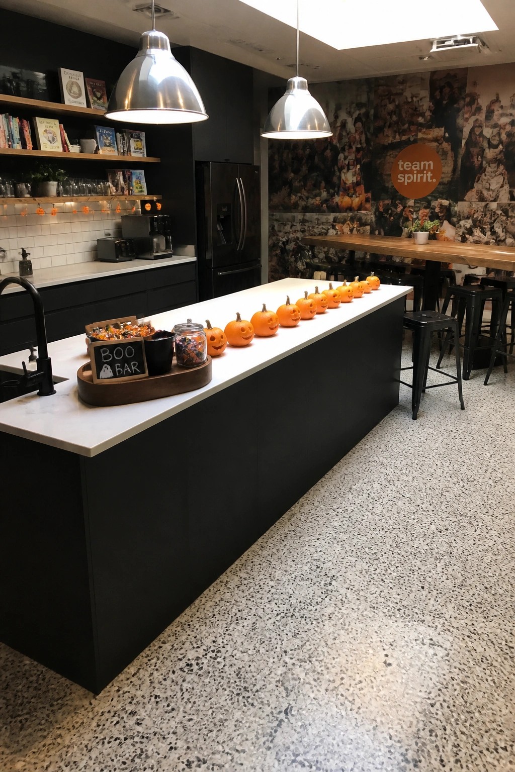 A modern office break room with a long row of small carved pumpkins placed along a black kitchen island, next to a wooden tray labeled 
