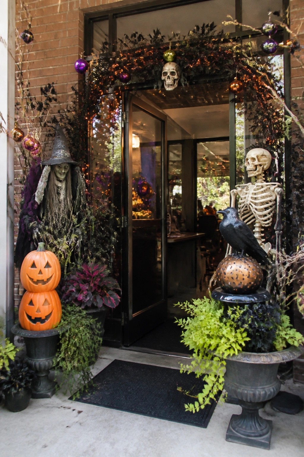 Office entrance decorated with a skeleton holding a crow, a standing witch figure, stacked jack-o-lanterns, and a skull garland around the doorway.