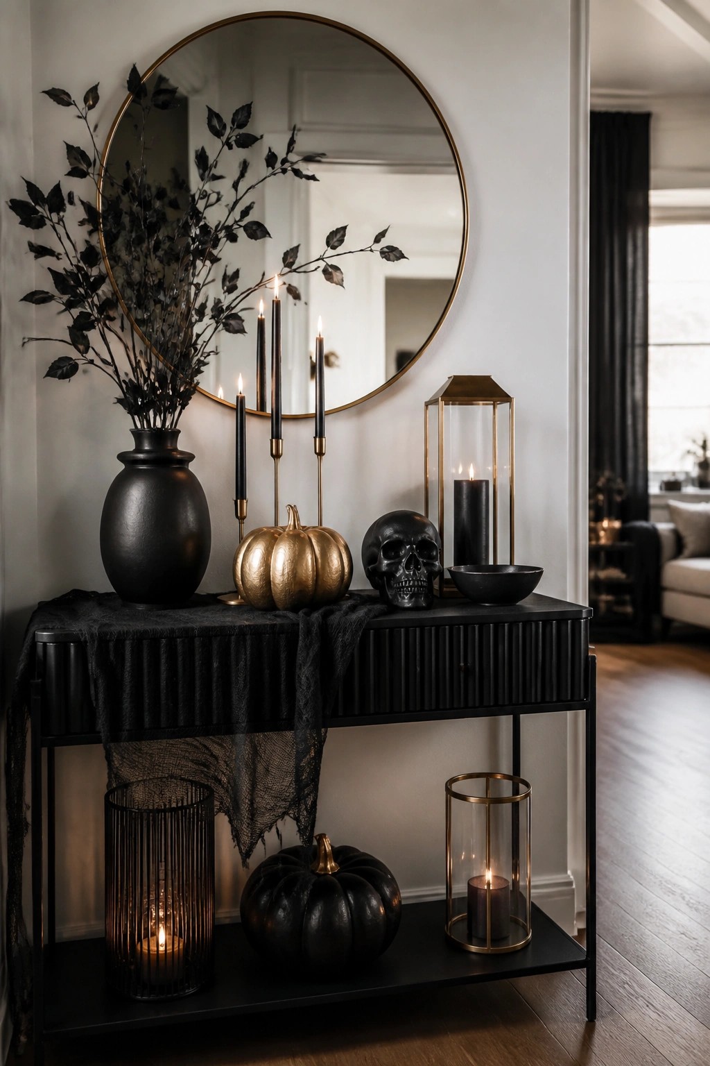 Entryway console table with a black skull, gold pumpkin, black vase holding dark branches, and several lit candles arranged under a round mirror.