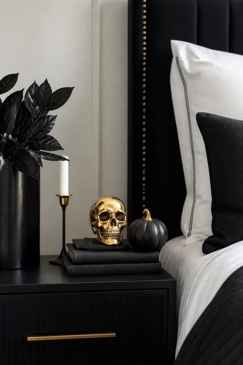 Bedroom nightstand with a gold skull resting on stacked black books, a black pumpkin, and a black vase holding dark leaves.