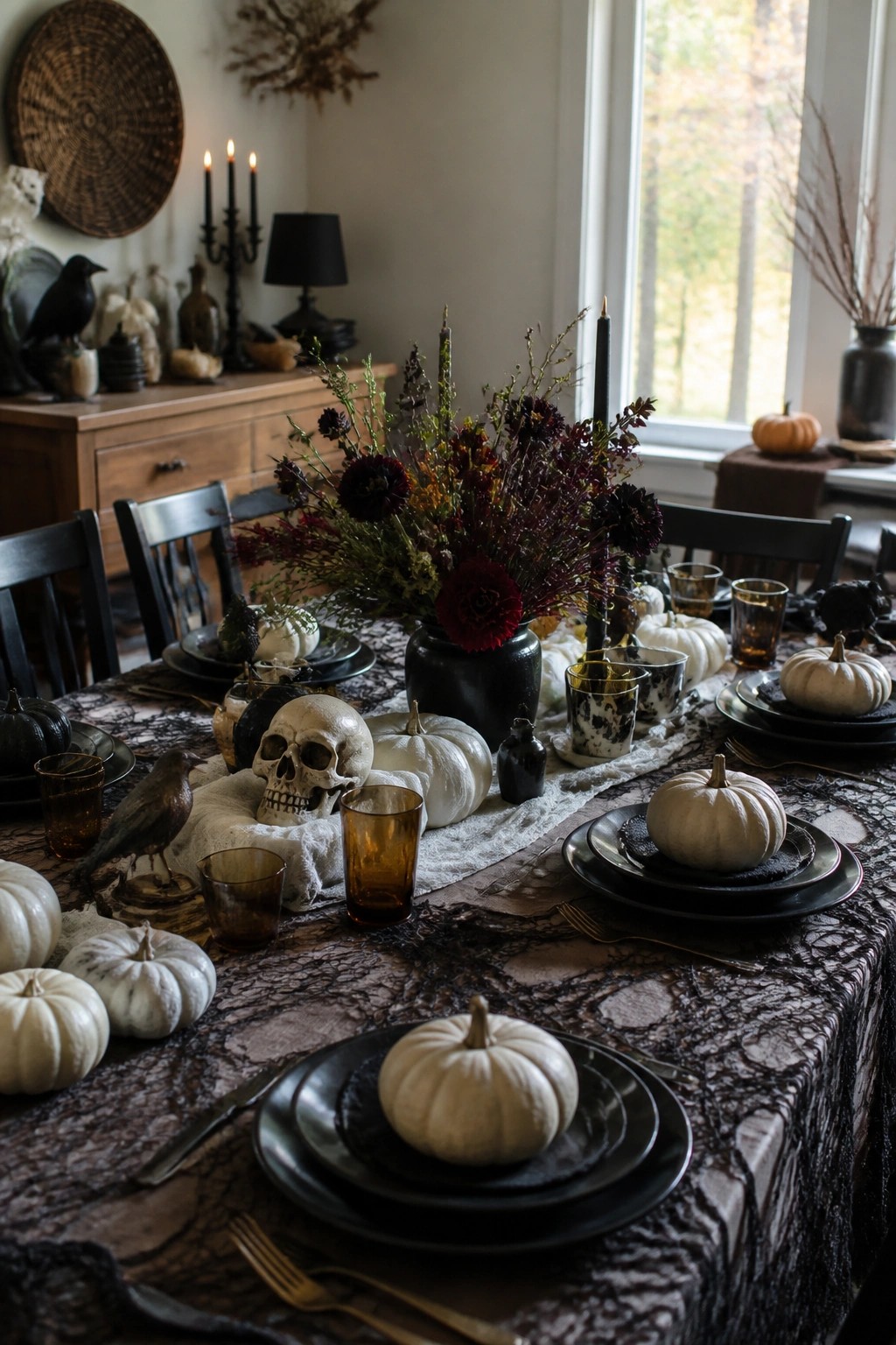 Dining table set for Halloween with a dark floral centerpiece, skulls, crows, white pumpkins, and black plates on a lace runner.