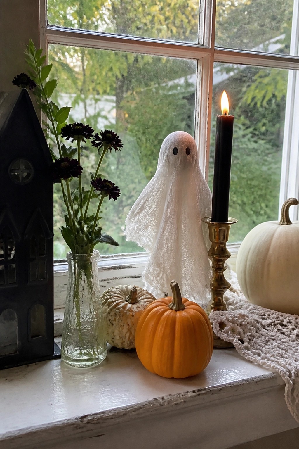 A white fabric ghost stands on a white windowsill beside an orange pumpkin, a white pumpkin, a lit black candle, and a glass vase of dark flowers.