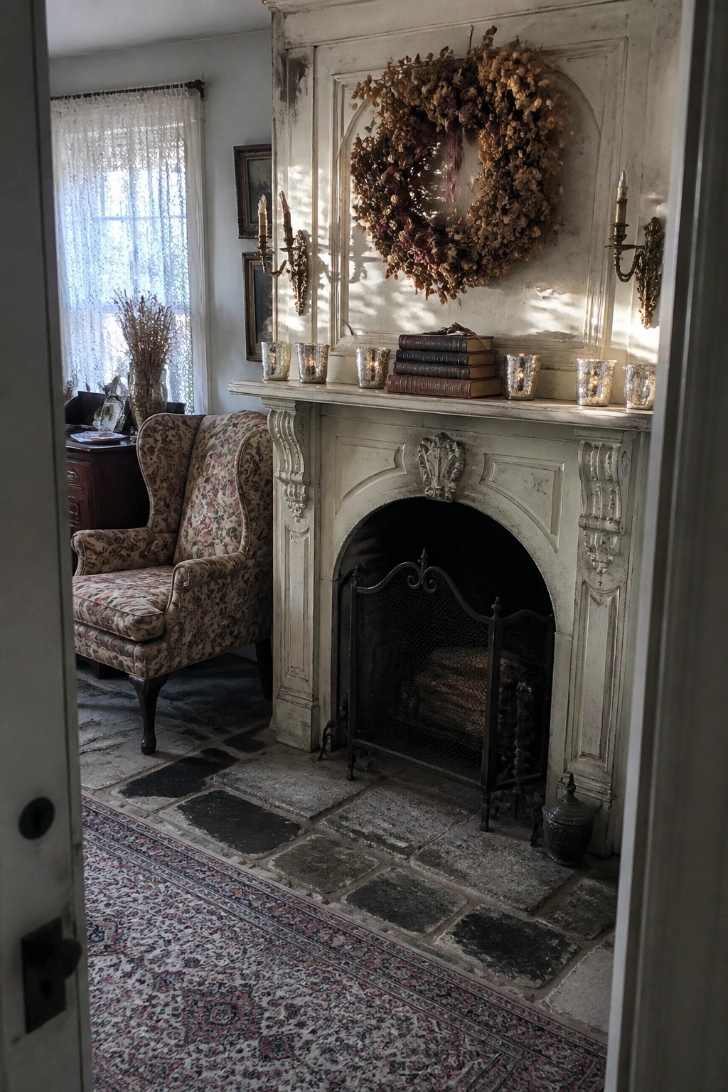 A white distressed fireplace mantel topped with a large dried floral wreath, stacked books, and mercury glass candle holders, viewed from a doorway in a room with a patterned armchair and stone floor.