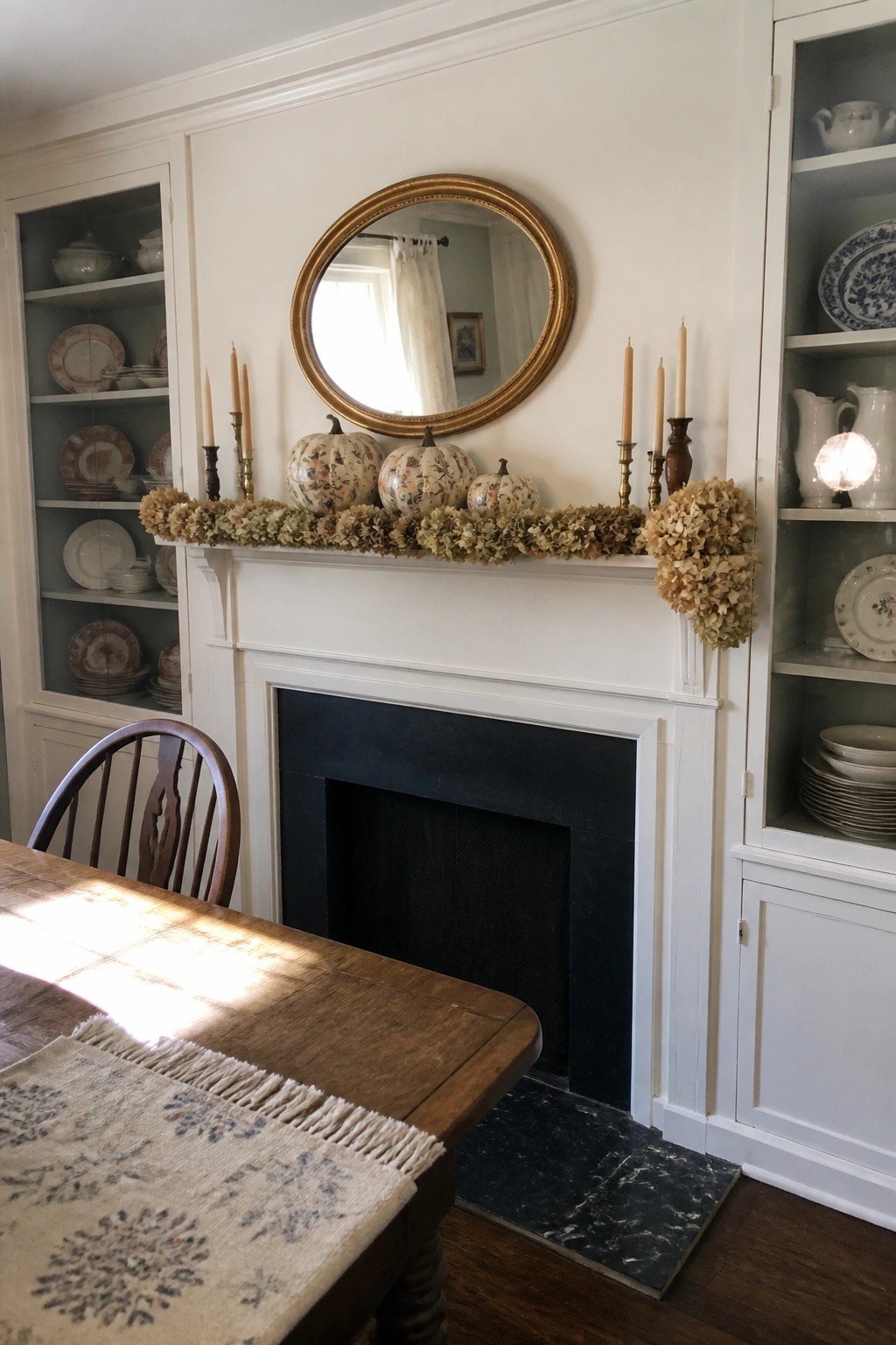 A white fireplace mantel with a dried hydrangea garland, three floral pumpkins, and several candles arranged below a round gold-framed mirror.