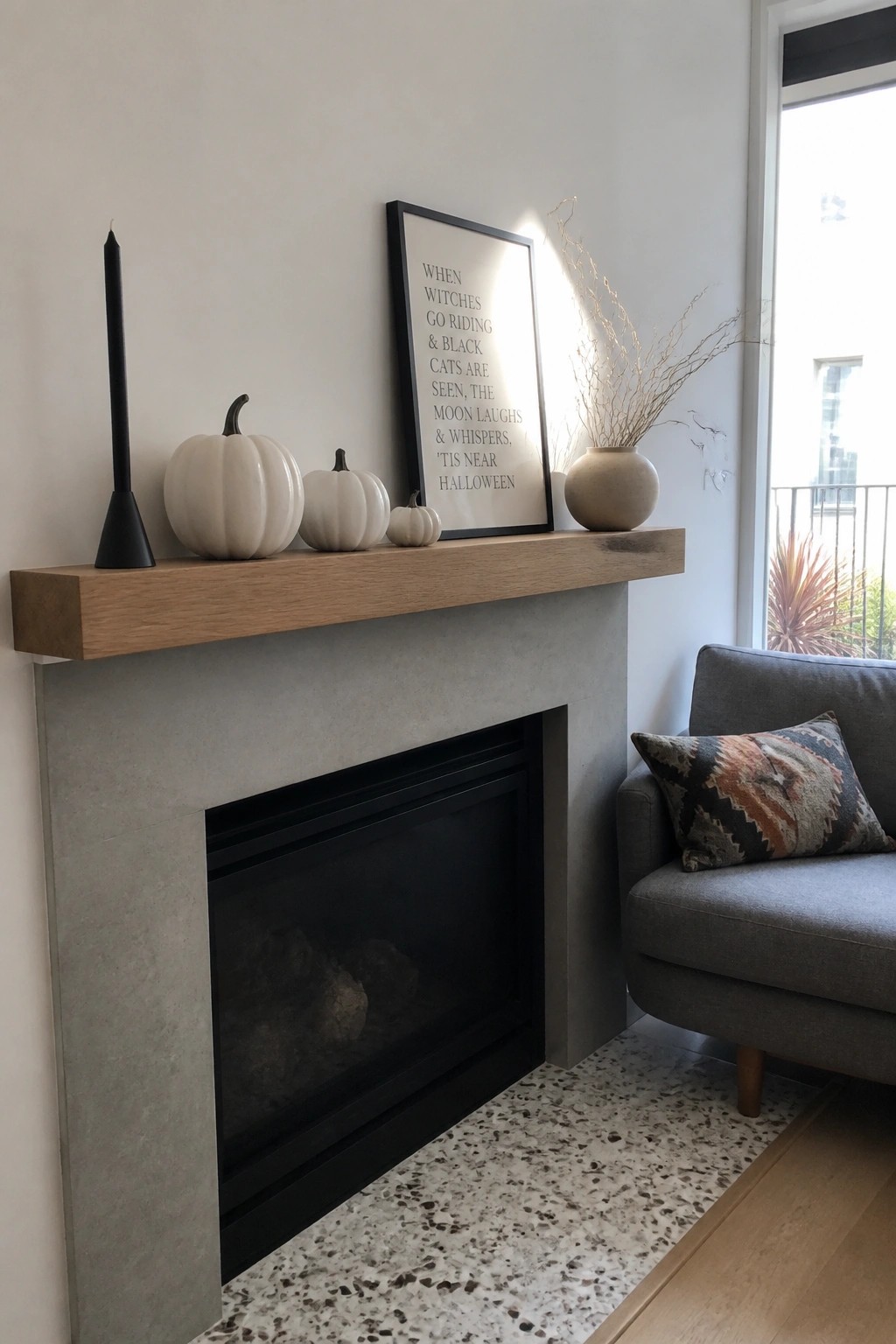 A concrete fireplace with a wooden mantel holding three white pumpkins, a black candle, a framed poem print, and a beige vase with dried branches.