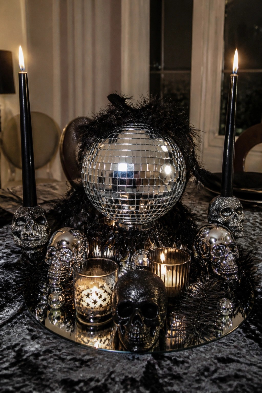 Disco ball centerpiece with glitter skulls, black feathers, and lit candles on mirror tray.