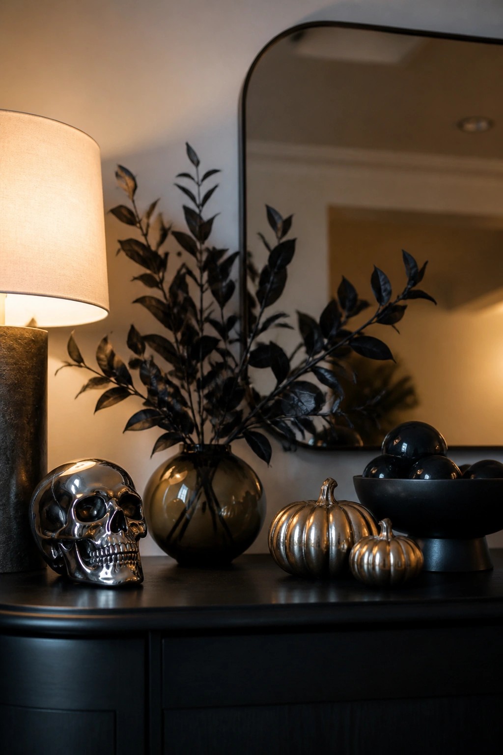 Dark console table with a chrome skull, two metallic pumpkins, a dark vase holding black branches, and a lamp next to a mirror.
