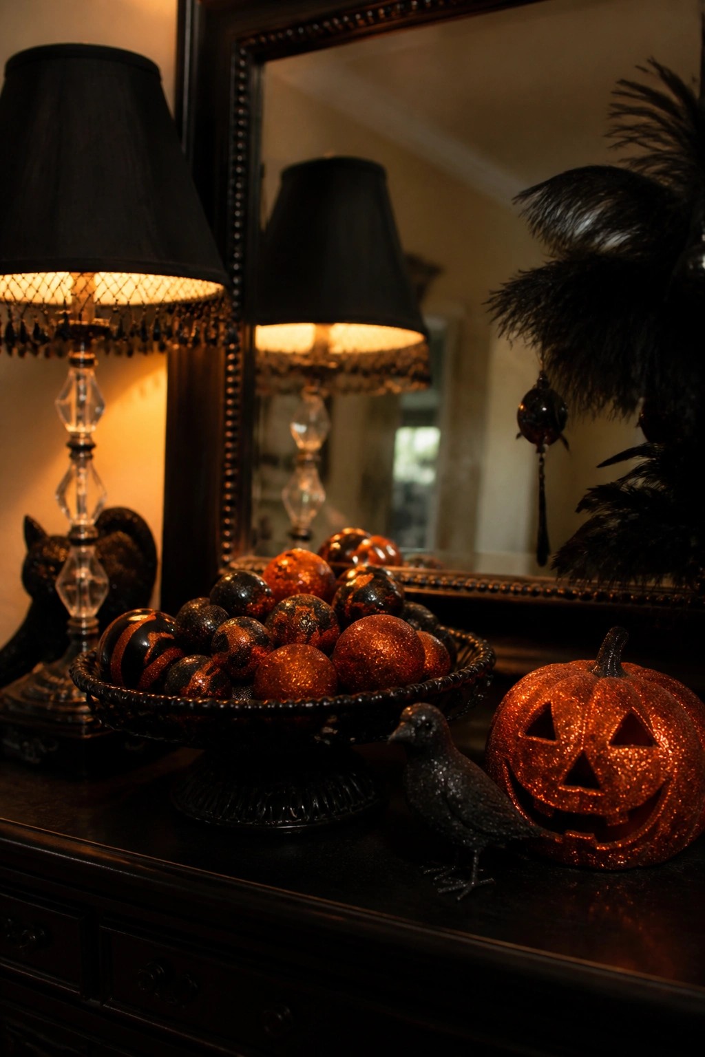 Dark wood console holding a glittery carved jack o lantern, a bowl of black and orange glitter orbs, a small black bird figurine, and black lamps with a mirror behind them.