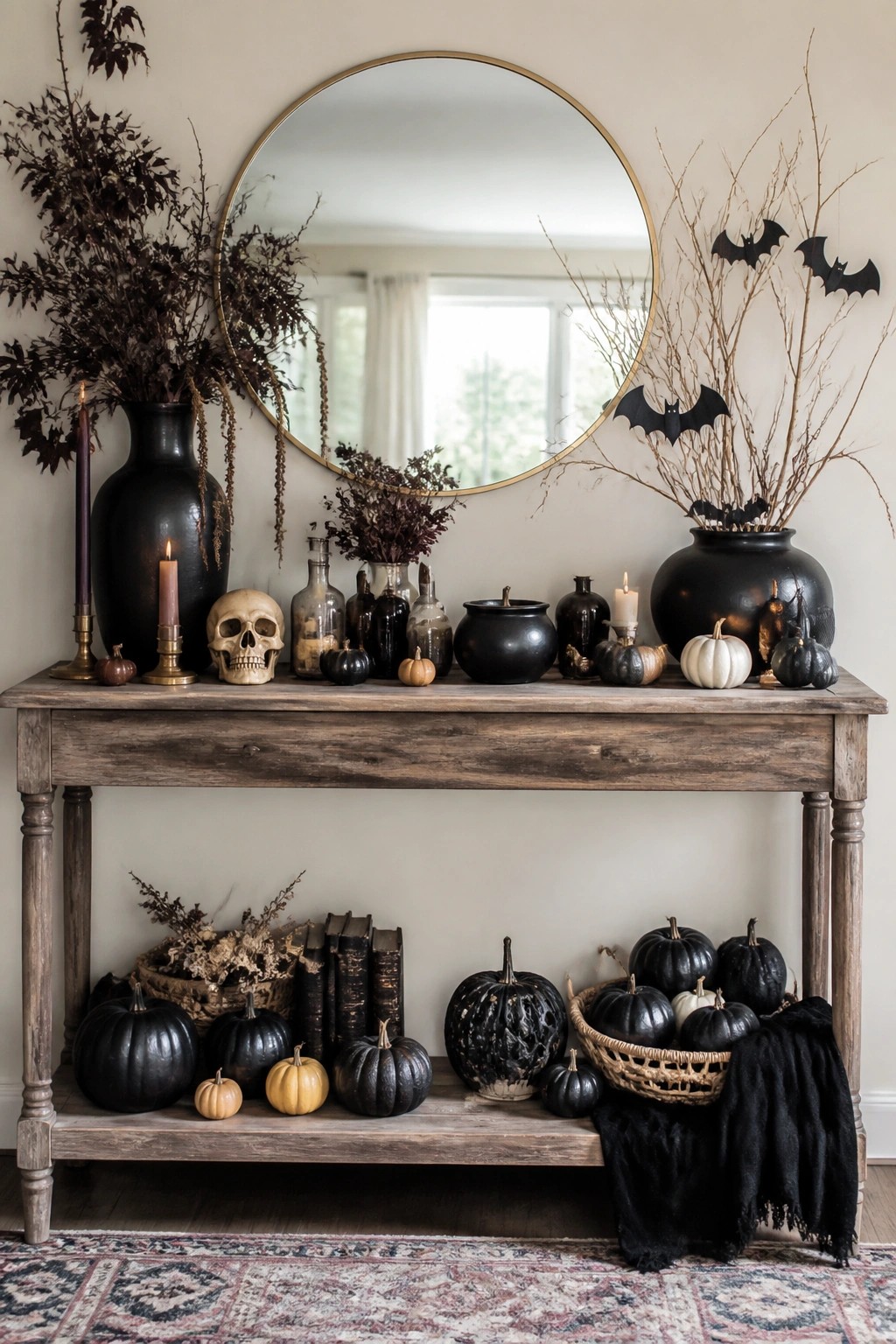 Entryway console table styled with black branches holding bat cutouts, a skull, dark vases, candles, and assorted pumpkins beneath a round mirror.