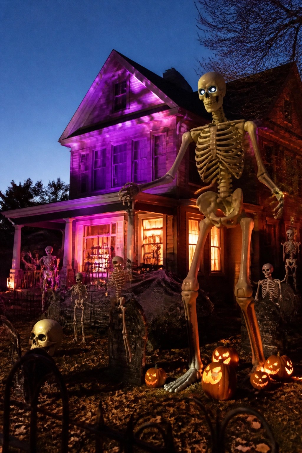 Tall skeleton decoration stands in a front yard with smaller skeletons, tombstones, and glowing pumpkins in front of a house at night.
