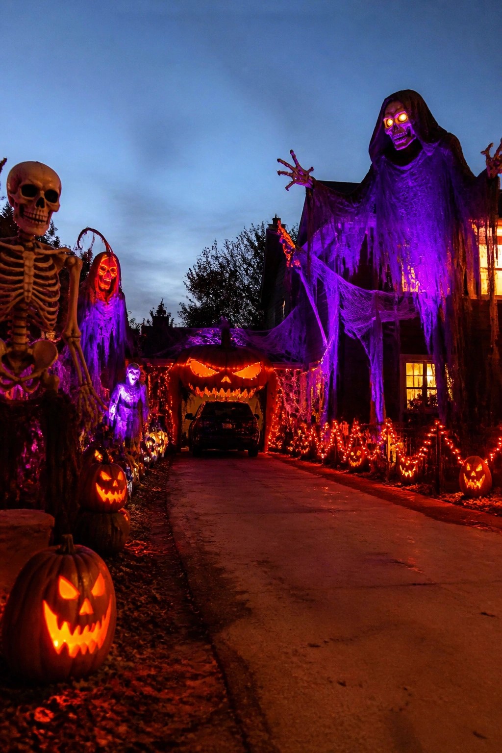 House exterior at night with a large purple-lit grim reaper figure hanging from the roof, spiderwebs stretched across the front, a giant carved pumpkin over the garage, and rows of smaller jack-o-lanterns along the driveway.