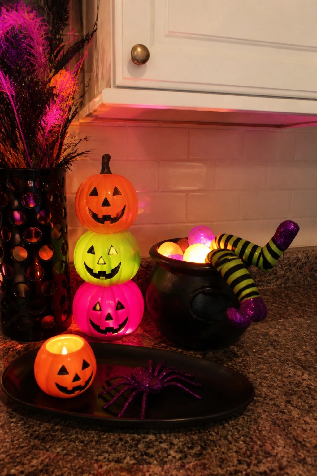 Kitchen counter with a stack of glowing orange, green, and pink jack o lanterns next to a black cauldron with striped witch legs and a small tray holding a lit pumpkin and purple spider.