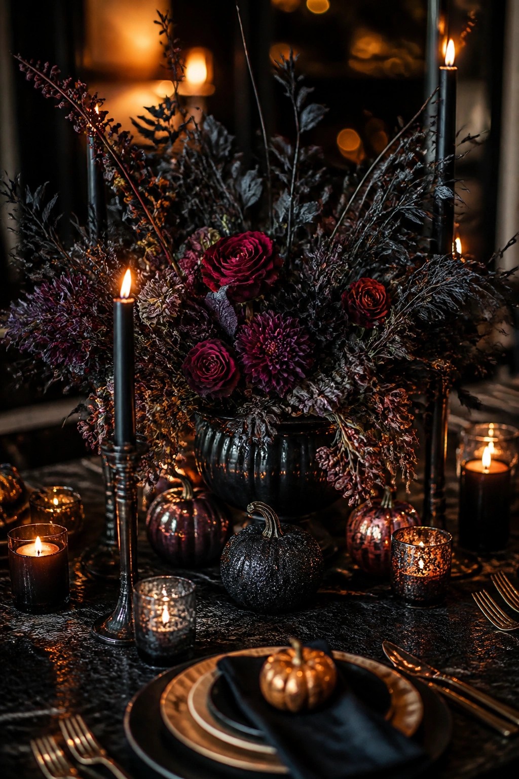 A dark dining table set with a large black vase of deep red and purple flowers, black taper candles, and small metallic and black pumpkins as the centerpiece.