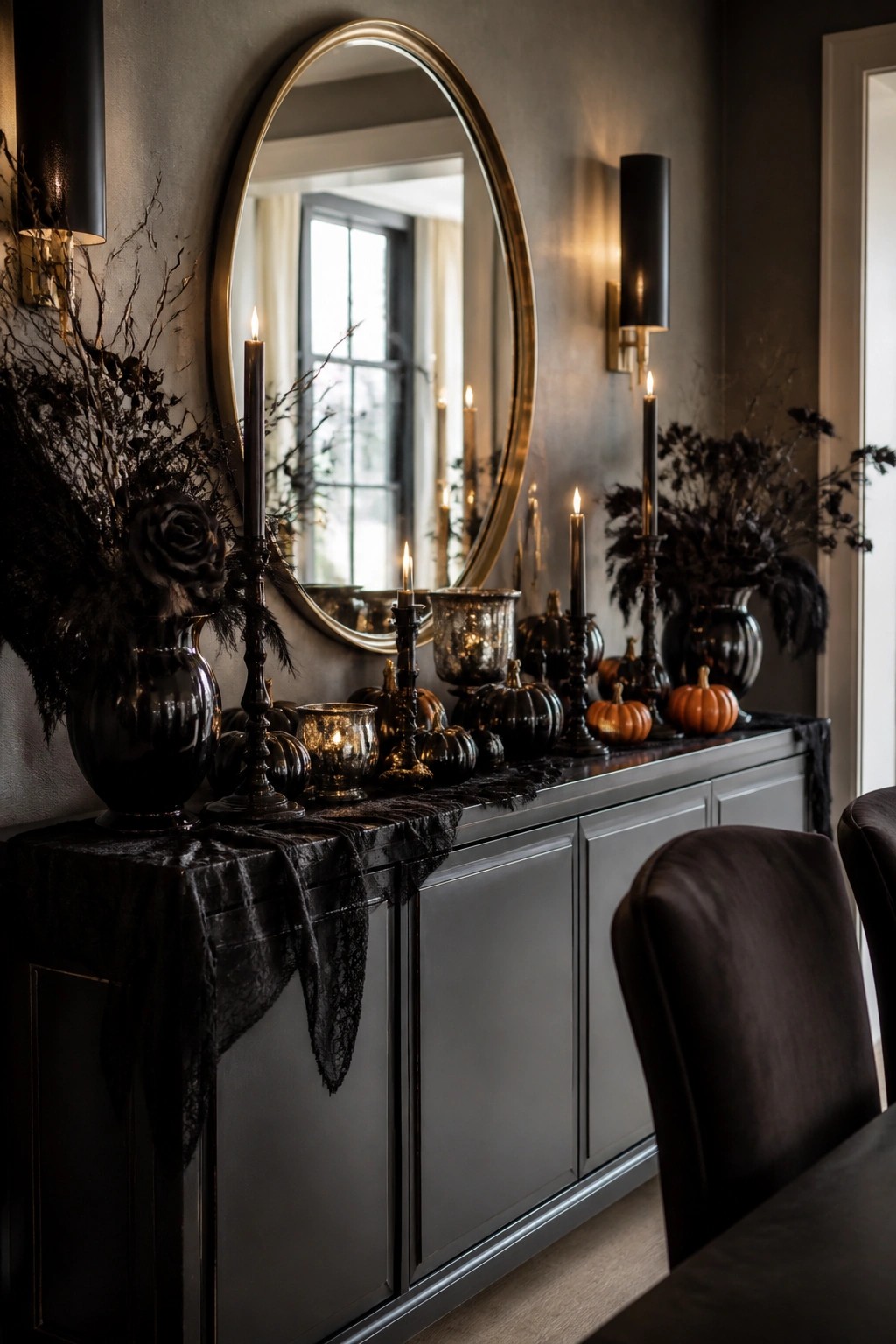 Dark console table decorated with black vases holding dark branches, black and orange pumpkins, lit candles, and a black lace runner beneath a large round mirror.