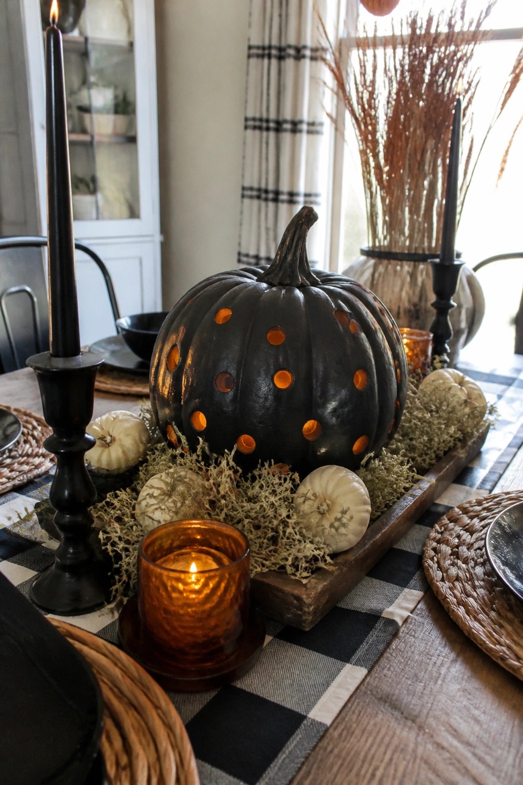 A black pumpkin with glowing circular cutouts sits on a wooden tray as the centerpiece of a checkered dining table, flanked by black candles and small white pumpkins.