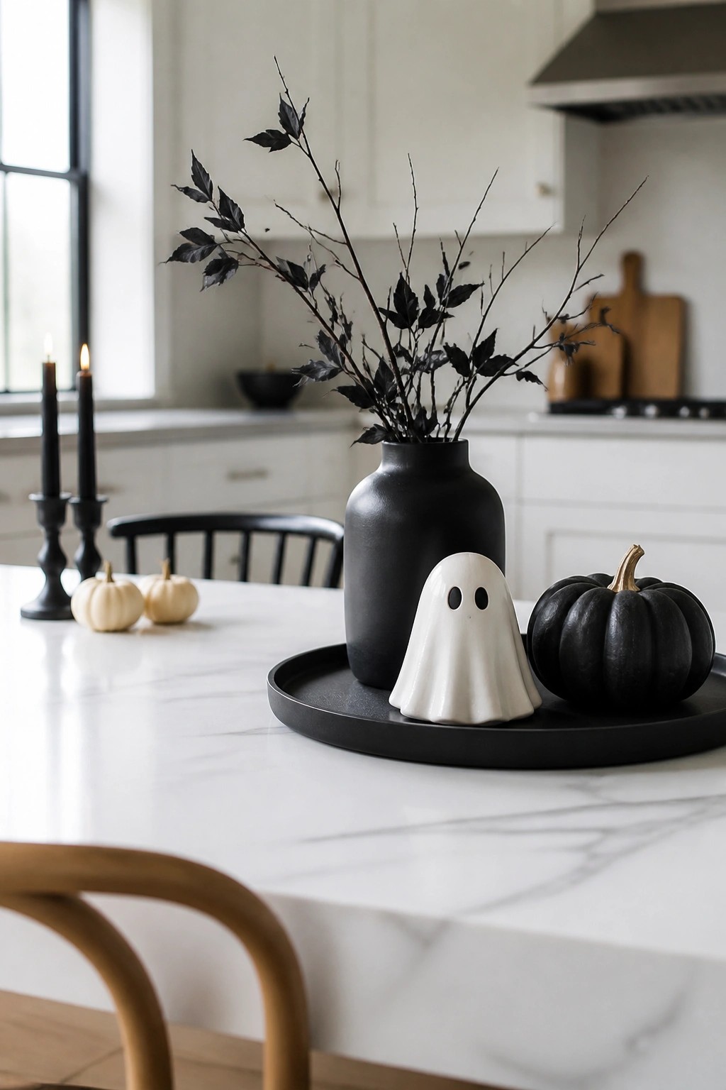 White ghost figurine on a black tray with a black vase of dark branches and a black pumpkin on a white marble kitchen counter.