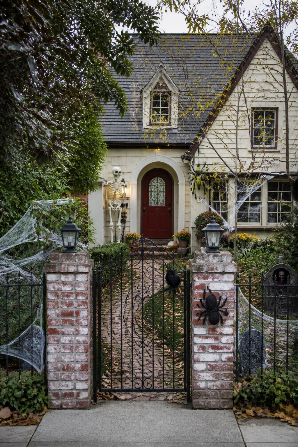 Front entrance with a skeleton standing beside a red door, spiderwebs on brick pillars, and a large spider on the gate.