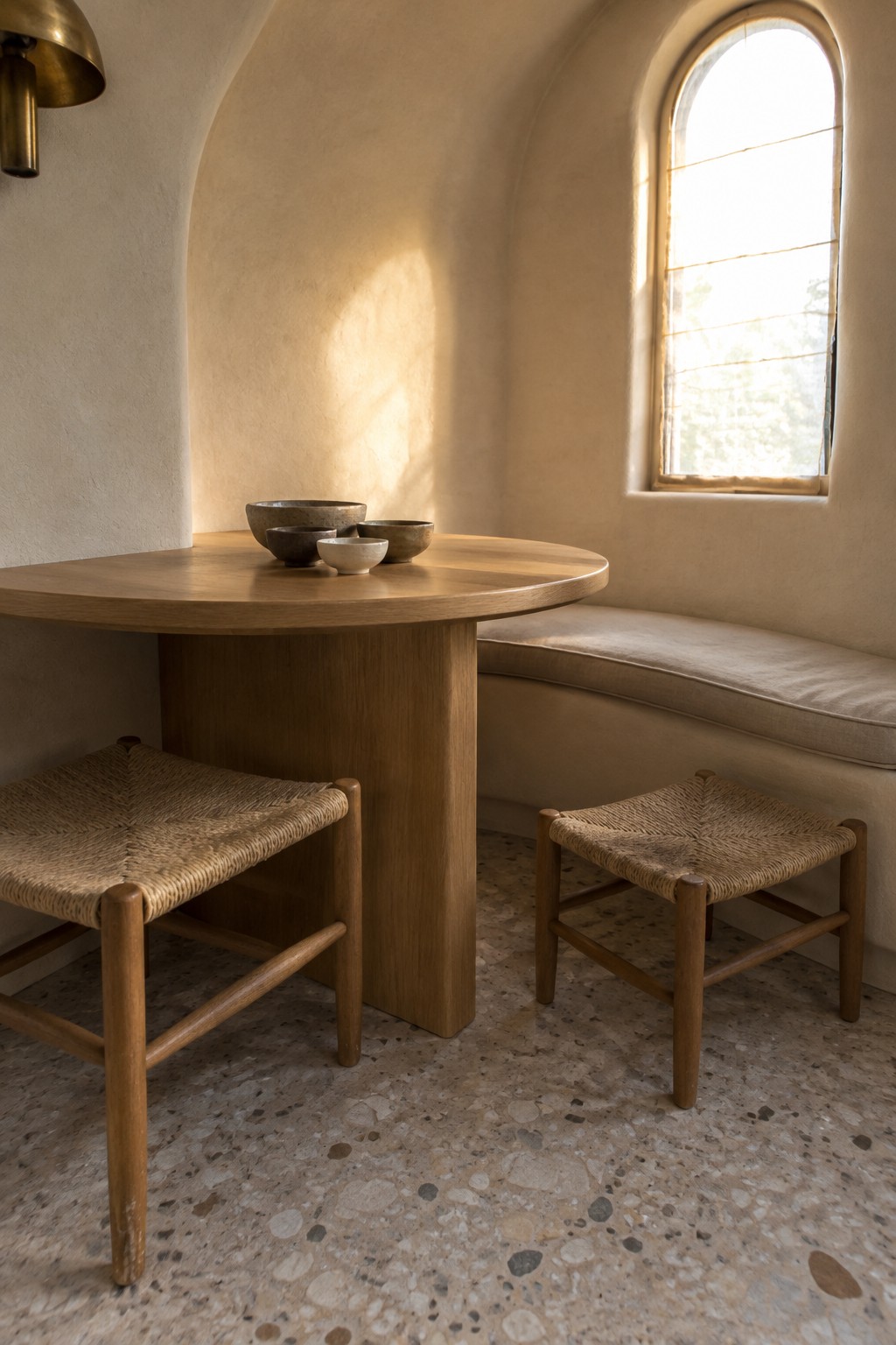 A round wooden pedestal table with ceramic bowls stands between two woven chairs and a built-in bench in a room with an arched window.
