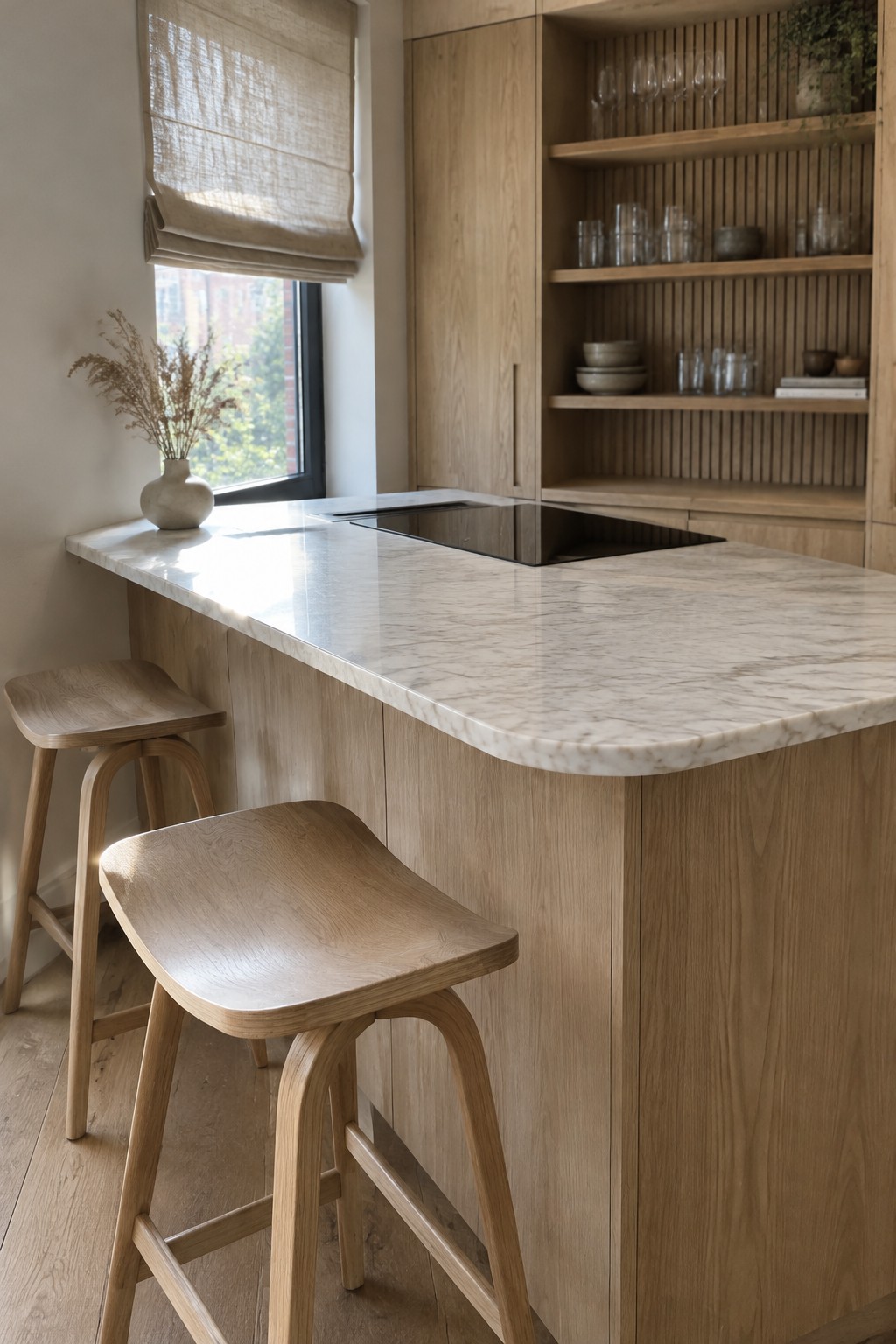 A light wood kitchen with a rounded marble island, black cooktop, wooden stools, and open shelves.