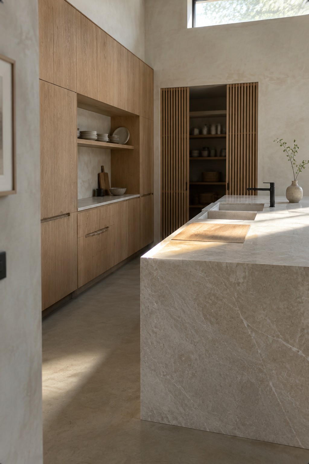 A kitchen with a pale stone island, oak cabinetry, two inset sinks, and a slatted pantry door.