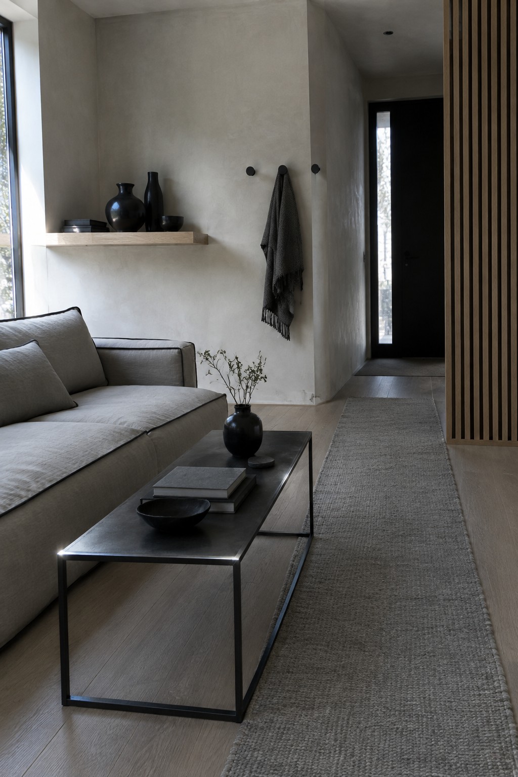 A narrow living room with a gray sofa, long textured runner, low black coffee table, and wood slat divider.