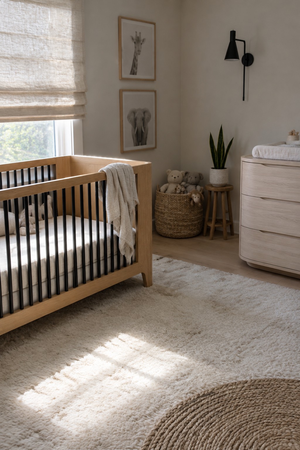 A nursery with a light wood crib with black rails, framed animal prints, a black wall lamp, and a woven basket of stuffed toys.