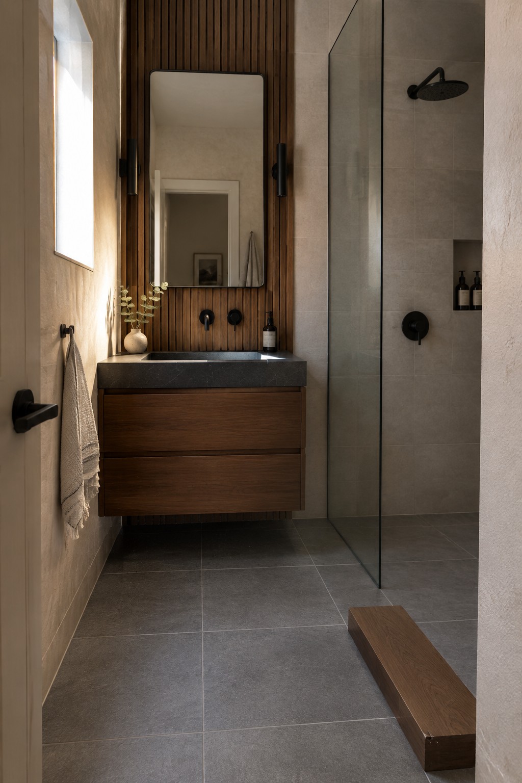 Warm greige stone tile in a bathroom beside vertical oak slats and a dark wood vanity.