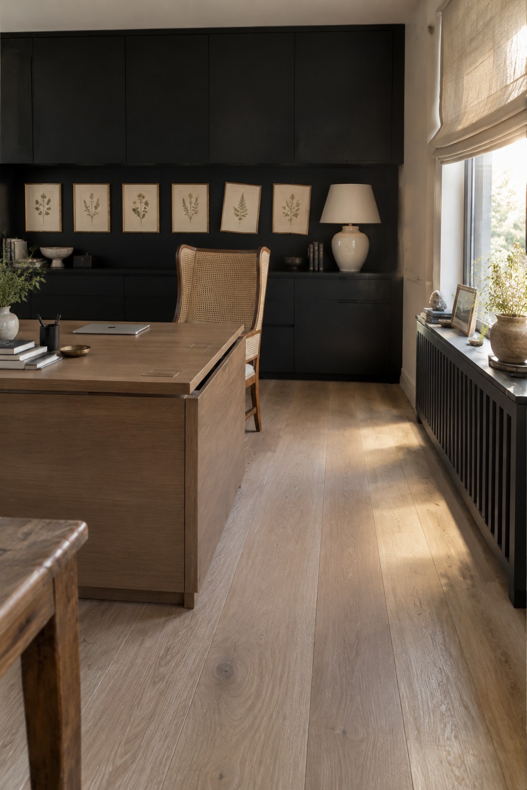 A wood desk sits in front of dark built-in cabinets in a home office with framed botanical prints and a window.